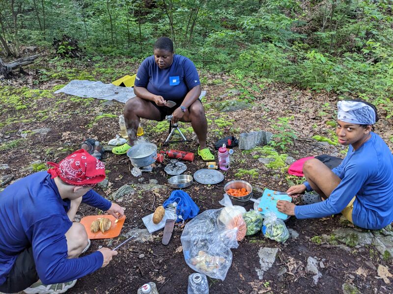 The image shows three people preparing food outdoors, possibly while camping. One person is sitting on a small stool, seemingly cooking over a portable stove. Another person is cutting vegetables on a cutting board, while the third person is also preparing food. There are various food items and cooking utensils scattered around them on the ground. The setting appears to be a wooded area.